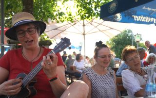 Frau mit Ukulele und Hut und Singende in einem Biergarten